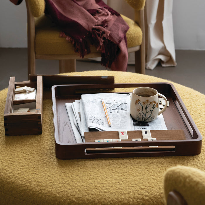 A dark brown wooden tray set on a yellow textured chair in a cozy room. The tray holds a folded newspaper, a white ceramic mug with a floral print, a pencil, and a small set of white tiles.