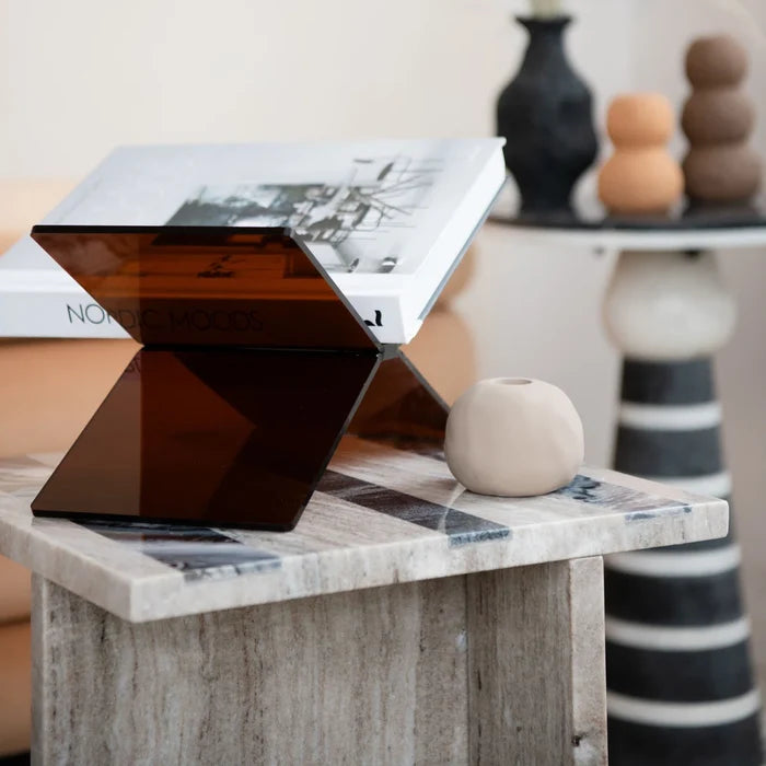 Dark tinted acrylic book holder displaying an open book on a wooden side table with minimalist decor in background