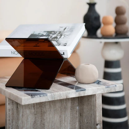 Dark tinted acrylic book holder displaying an open book on a wooden side table with minimalist decor in background