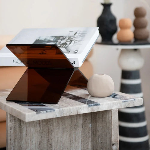Dark tinted acrylic book holder displaying an open book on a wooden side table with minimalist decor in background
