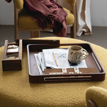 A dark brown wooden tray set on a yellow textured chair in a cozy room. The tray holds a folded newspaper, a white ceramic mug with a floral print, a pencil, and a small set of white tiles.