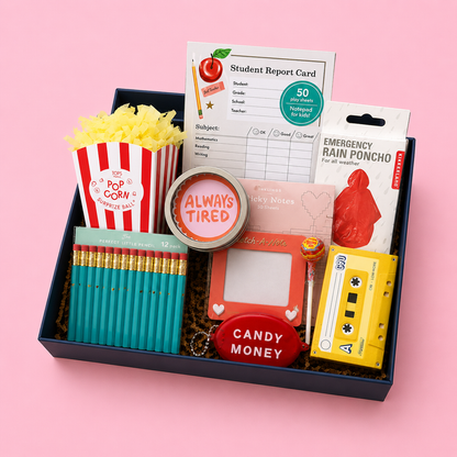 A curated school-themed gift box set in a navy box, featuring a red and white striped popcorn bucket with yellow paper, teal pencils, a round tin reading 'ALWAYS TIRED', a pink notepad, a yellow cassette tape notebook, a red candy money coin pouch, and other school supplies on a pink background.