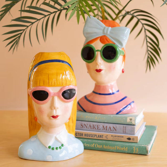 Two colorful ceramic lady vases wearing sunglasses, one with a bow and one with a headband, displayed with books and plant leaves.