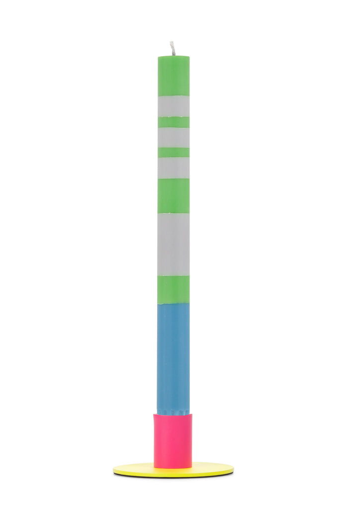 Tall colorful candle with green, white, blue, and pink sections on a yellow metal holder against a white background