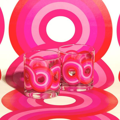 Two clear rocks glasses with bold pink and red retro swirl patterns on a matching colorful backdrop